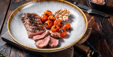 Roasted Lamb tenderloin meat in plate with grilled tomato and garlic, mutton sirloin fillet steak. Dark background. Top view.